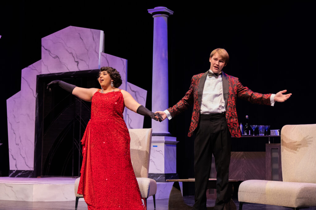 two actors on stage hold hands, the woman is in a red dress and the man a red and back tux. The stand in front of an purple pill and archway.