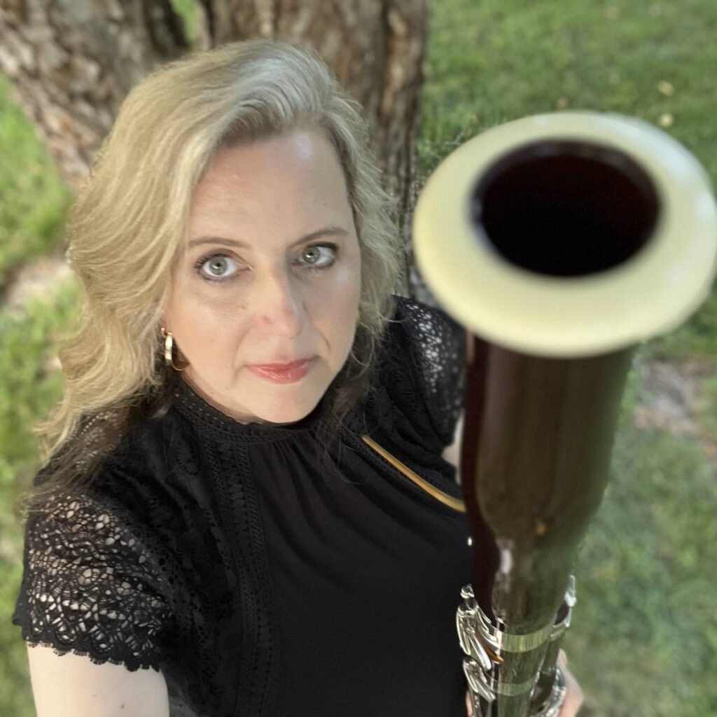 a woman in a black blouse stands in front of a tree trunk holding a bassoon